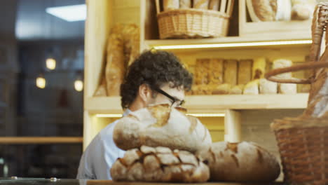 Hombre-Apuesto,-Vendedor-De-Panadería-Con-Gafas,-Trayendo-Pan-Fresco-En-El-Mostrador-Por-La-Mañana-Y-Oliéndolo