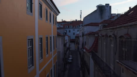 Backwards-flying-drone-camera-between-buildings-in-evening-downtown.-Elevated-view-of-old-houses-lining-narrow-streets.-Lisbon,-capital-of-Portugal.