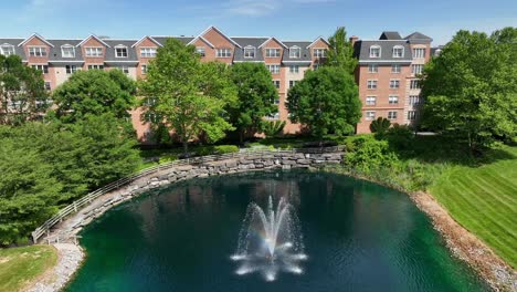 Drone-view-of-an-apartment-complex-behind-a-park-like-setting-with-a-small-lake-with-a-fountain