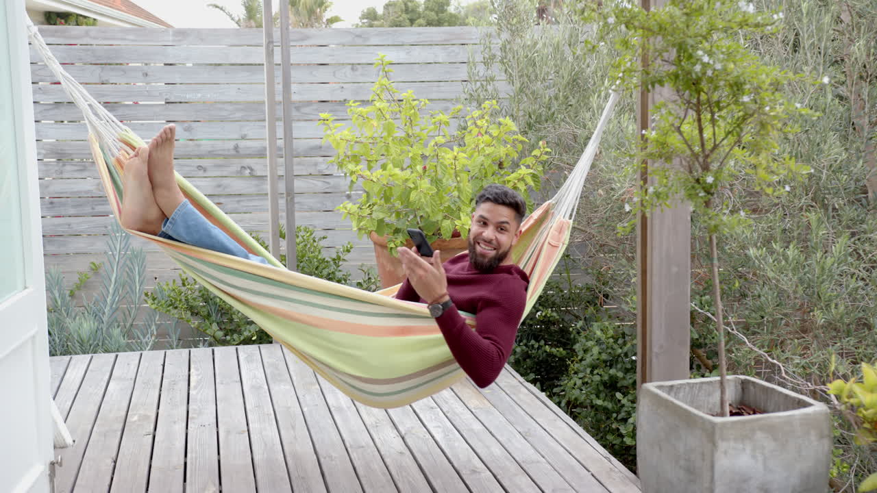 Relaxing in hammock, man using smartphone and smiling on wooden deck