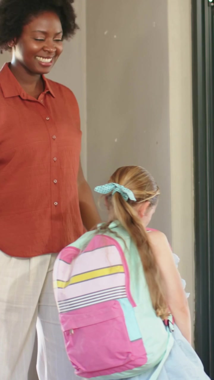 Vertical video: Greeting adult guiding girl with pastel backpack through glass door to enter room