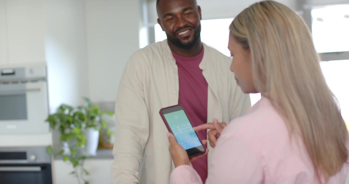 African American couple in kitchen woman tapping maroon phone, man noticing approaching, copy space