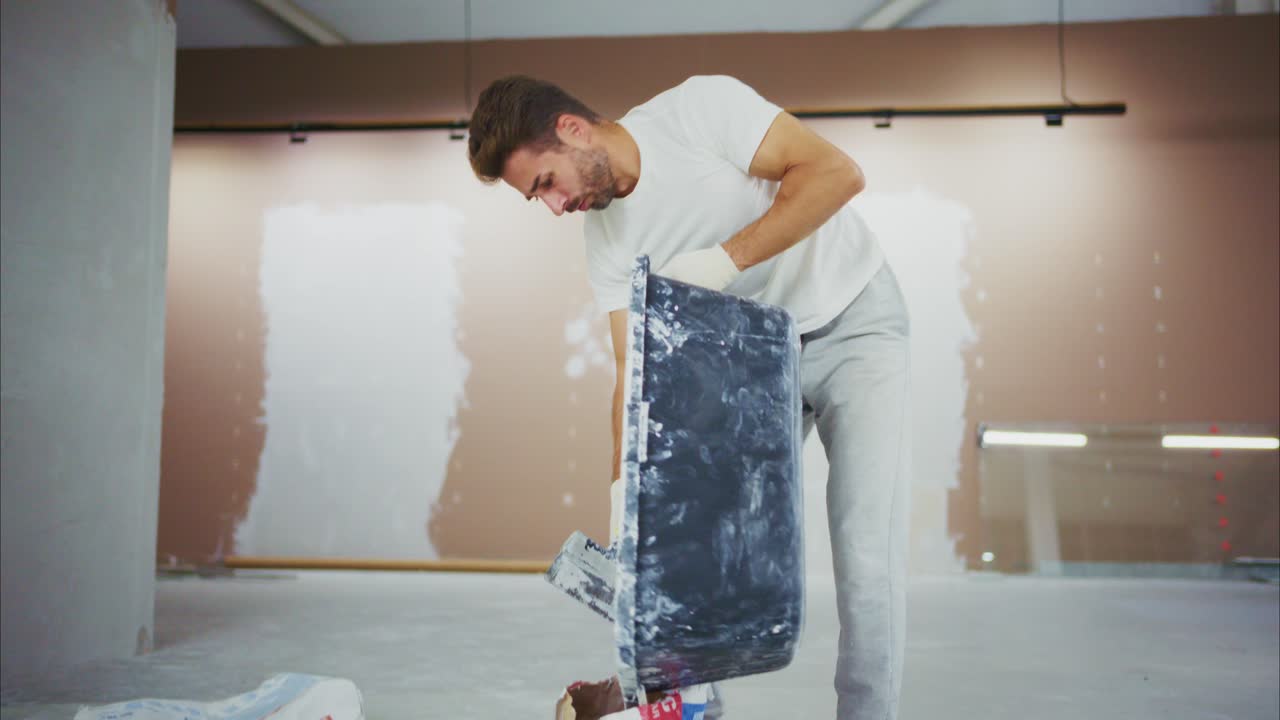 A diligent worker meticulously handles a tub of plaster, focused on his task within a partially renovated interior space, showcasing dedication to craftsmanship and detail