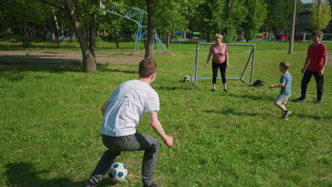 familia involucrada en un juego de fútbol al aire libre, el niño de blanco mientras dribla la pelota hacia la meta, sus hermanos intentan detenerlo, él dispara a la pelota, la madre rápidamente patea la pelota