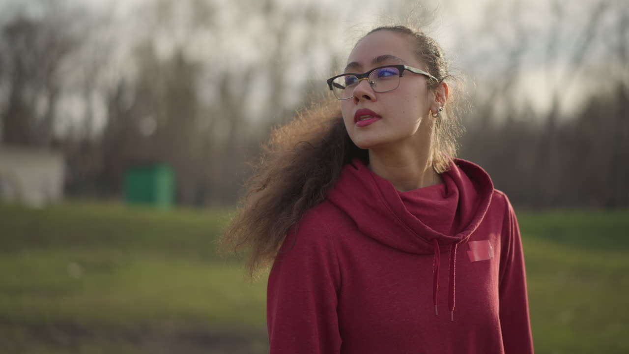Girl In Open Field Turns Head Thoughtfully, Teenage Girl With Glasses Surveys Expansive Landscape Curiously, Young Girl With Tousled Hair And Glasses Examines Open Area With Wonder