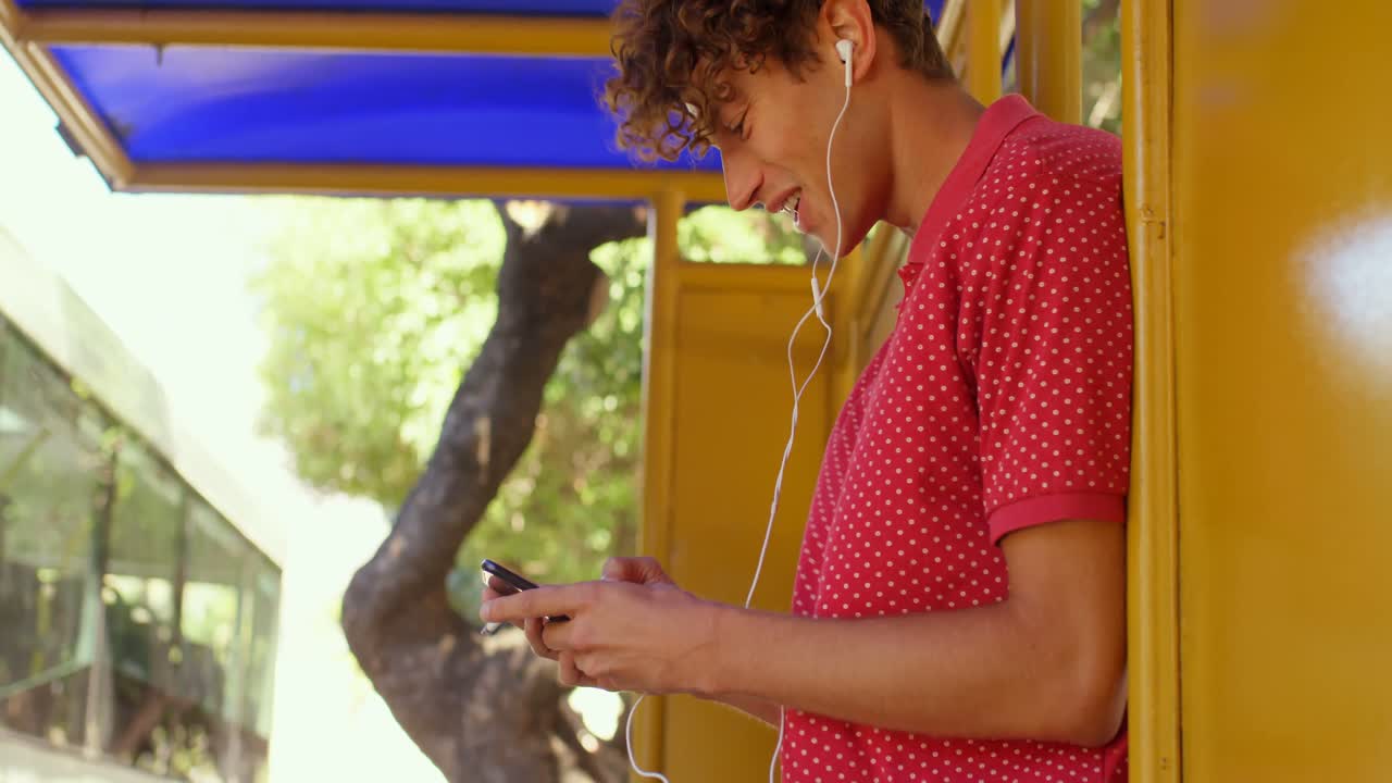 hombre escuchando música en el teléfono móvil mientras está de pie en la parada de autobús 4k