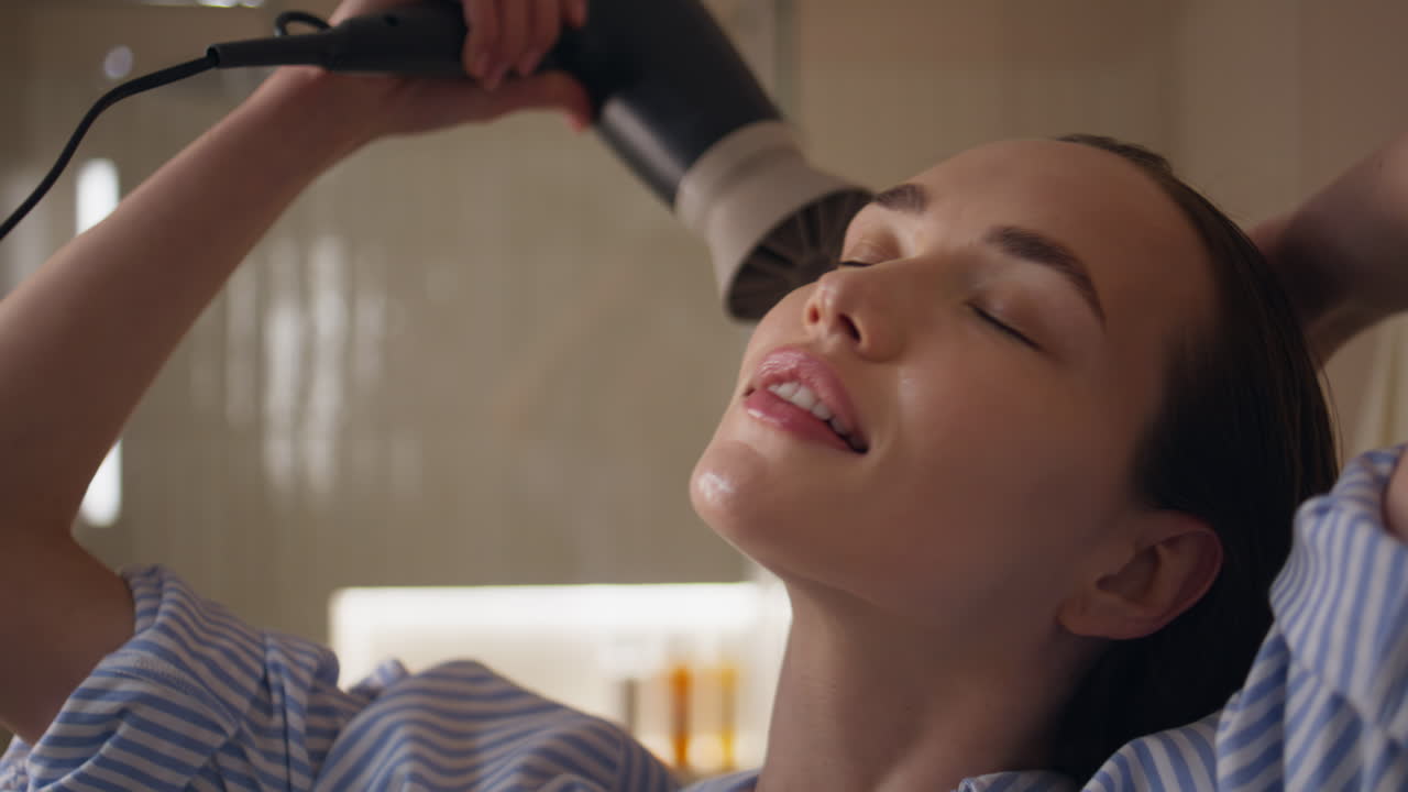 Nahaufnahme eines Mädchens, das morgens zu Hause die Haare mit einem Föhn stylt. Tägliche Haarpflegeroutine.