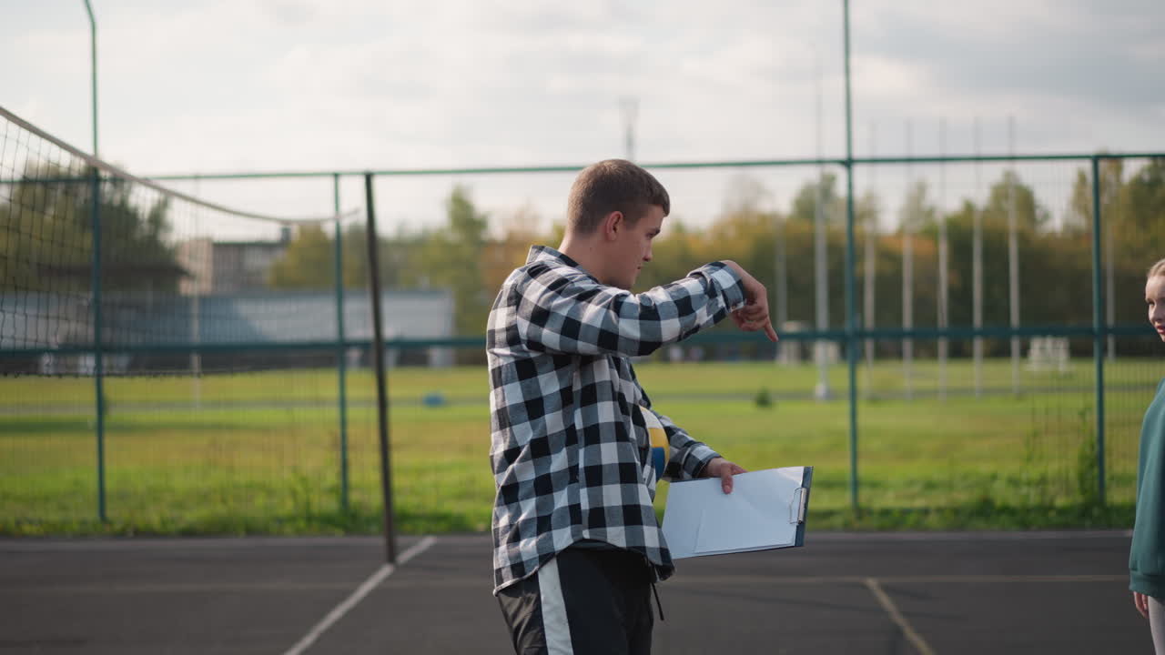 Volleyball coach with ball under arm and record book instructing female students to walk around court during practice session in outdoor environment, giving directions to beginners