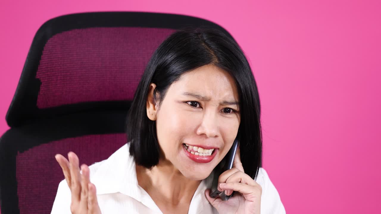 Shocked, frustrated woman in white shirt on phone, seated against a pink office background