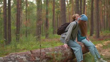 two tired hikers rest on fallen tree in peaceful forest, one wearing blue bandana lifting leg and gazing at companion, who rests hand on tree trunk, surrounded by tall trees and lush greenery