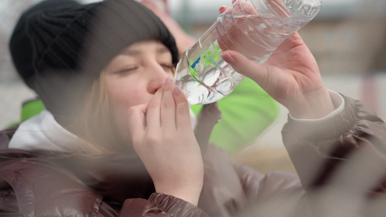 골대 옆에서 플라스틱 물병으로 물을 마시는 백인 여성, 검은색 비니와 갈색 코트를 입고 물을 벌컥벌컥 마시며 갈증을 해소하고 있으며, 밝은 색 재킷을 입은 동료가 뒤에 있고, 추운 날 휴식을 취하는 솔직한 모습