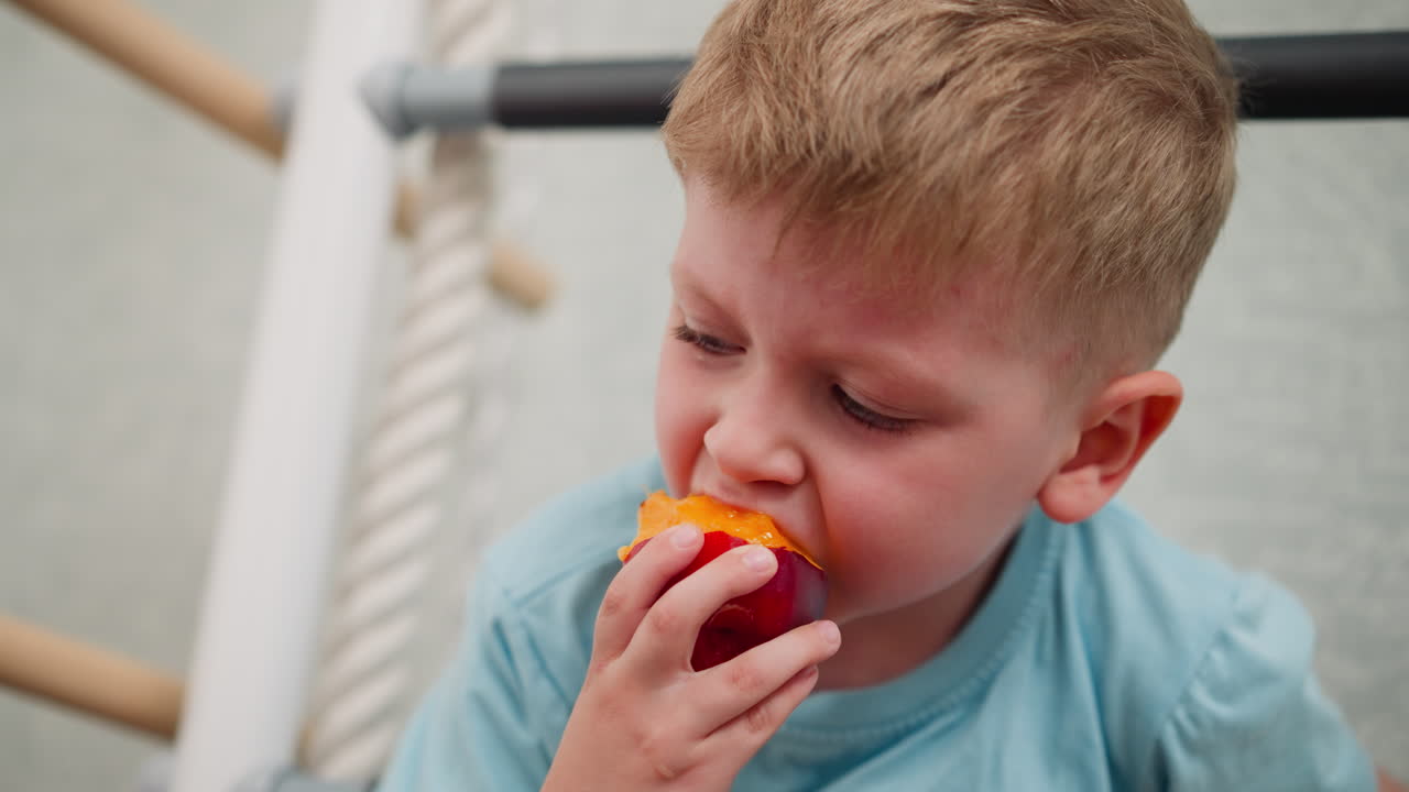 Young boy in blue top hanging on indoor ladder biting juicy fleshy mango with playful expression enjoying fresh fruit snack at home in brightly lit cozy room focused on savoring sweet tropical flavor