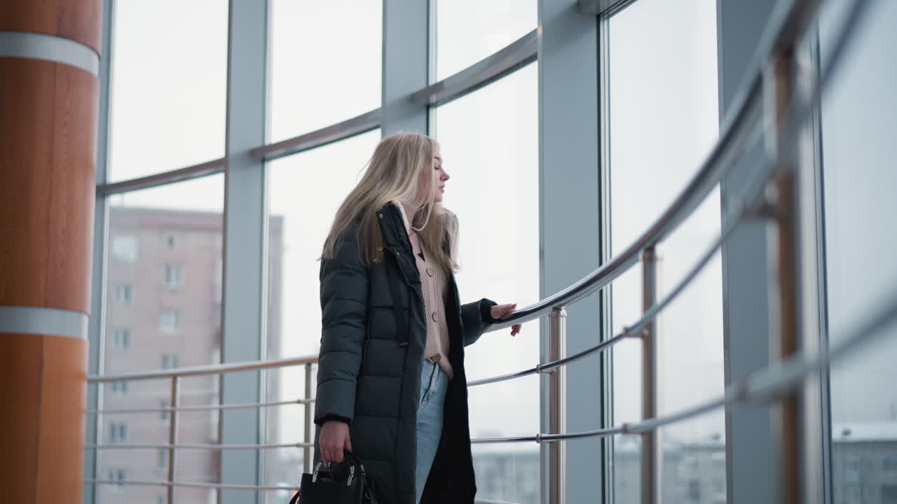 Lady standing near iron railing, holding black handbag, gazing out through glass with cityscape and office building background, offering a peaceful moment in a modern urban environment
