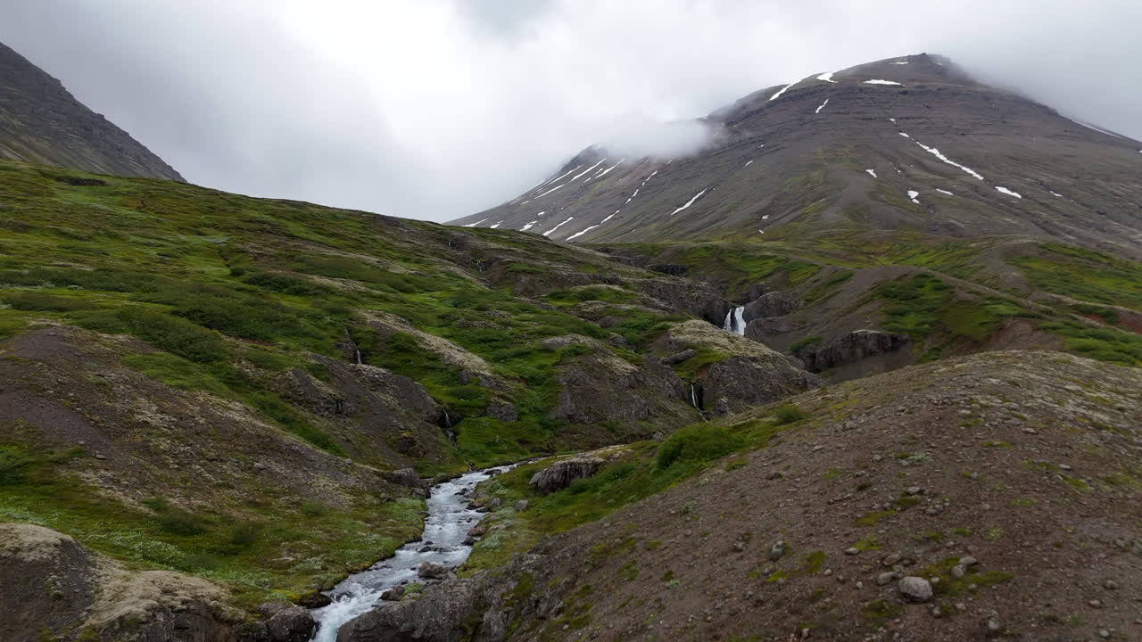 Vista aérea de drone de uma paisagem nublada e mística perto de Útsýnispallur Grænafell, com montanhas escarpadas, cordilheiras nebulosas e riachos em cascata moldando as terras altas selvagens da Islândia
