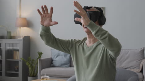 homme senior utilisant des lunettes de casque de réalité virtuelle et déplaçant ses mains tout en étant assis sur une chaise dans un salon moderne