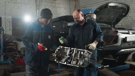 two mechanics in blue uniforms, one passing a tool to another while working on an engine in a well-equipped workshop, background shows a car and various mechanical tools