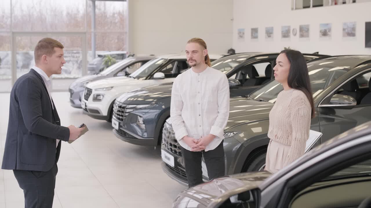 un joven vendedor de coches mostrando a una pareja de jóvenes un nuevo automóvil en el salón de concesionarios.