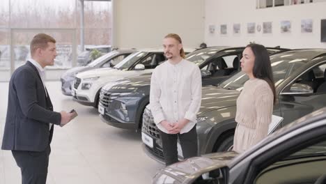 young car salesman showing to young couple new automobile at dealership salon.