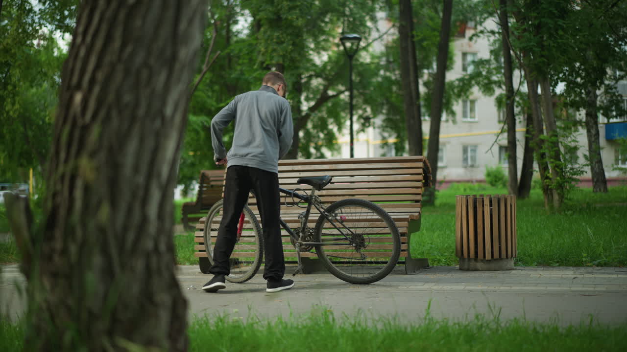 어린 소년은 공원으로 자전거를 타고 나무 벤치에 앉아 자전거의 바를 만져서 자세히 조사합니다. 초록색 나무, 풀, 그리고 배경의 주거 건물로 둘러싸여 있습니다.