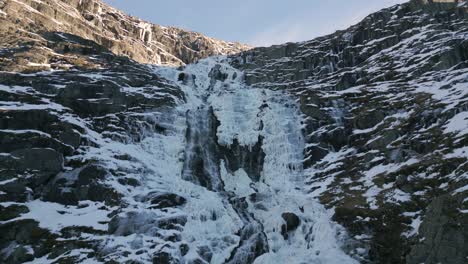 frozen waterfall cascading down rugged cliffs in norway, sunlight glancing off the icy surface