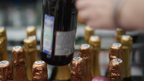 a hand wearing beaded bracelets selecting a wine bottle with gold foil from a shelf in a store. shot with a handheld camera, capturing the moment of choosing a wine bottle among many