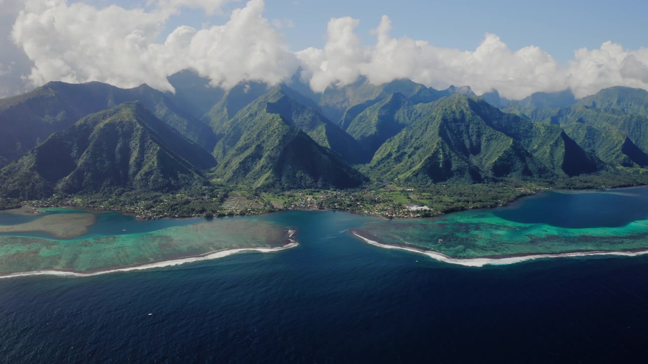 Aerial drone shot over Teahupoo valley, Tahiti, bright sky above twin turquoise reef passes framed by green mountains and scattered clouds.