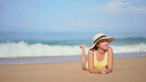 asian girl wearing yellow swimsuit and big hat lying on beach smiling looking at camera