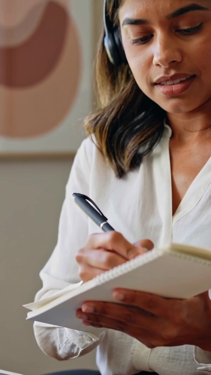 Vertical video: Pressing laptop keys woman in white top at home writing notepad and donning headset