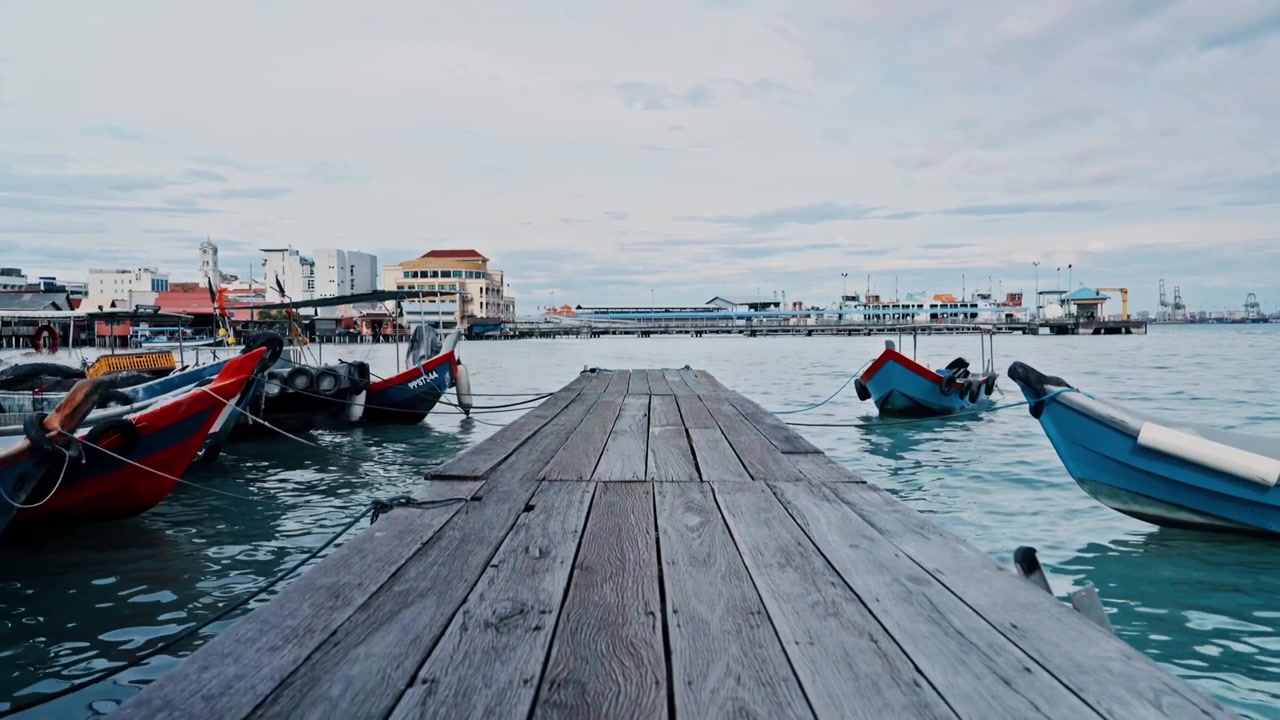 Et lavt perspektiv af en rustik træmole, der fører hen mod fortøjede traditionelle blå og røde fiskerbåde ved en klanmole, med Penang færgeterminal synlig i det fjerne.