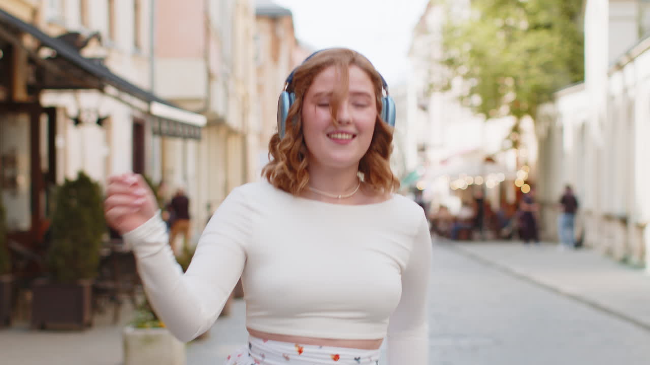 feliz mujer pelirroja sonriente en auriculares inalámbricos escuchando música bailando al aire libre en la calle de la ciudad