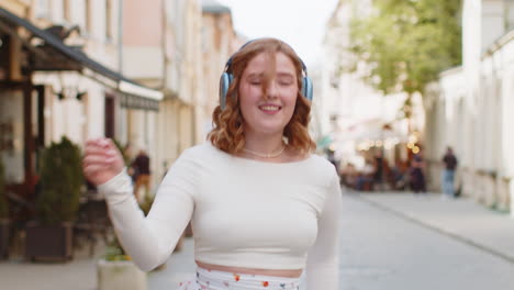 happy smiling redhead woman in wireless headphones listening music dancing outdoors in city street