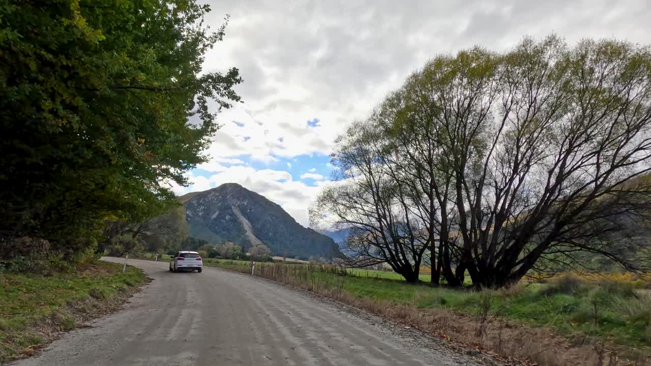 Vehicle travels on rural gravel road through lush trees, mountains, and cloudy sky, steady camera