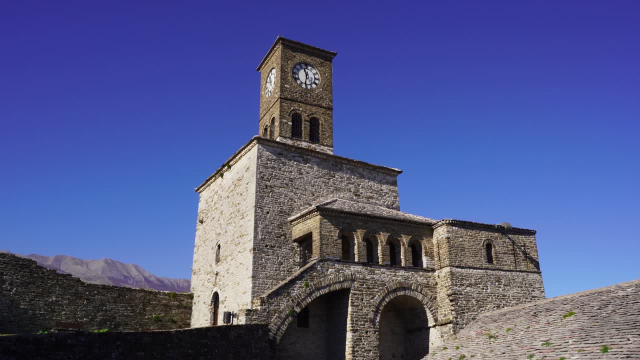 torre de reloj medieval rodeada de paredes de piedra sobre fondo de cielo azul brillante en gjirokastra