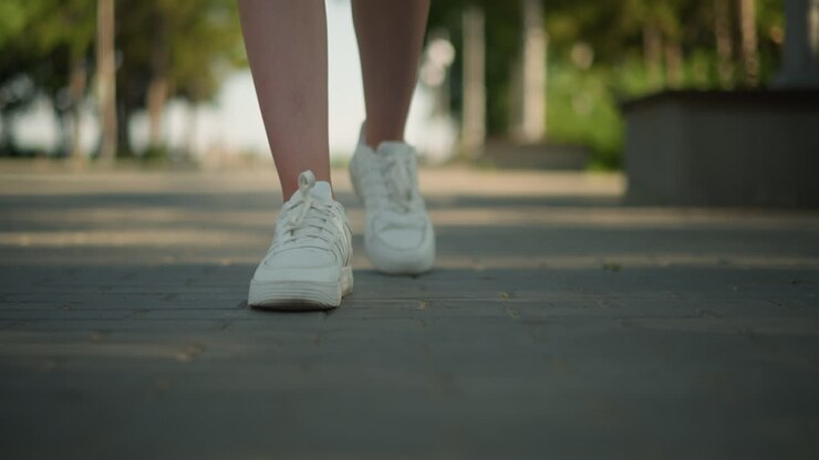 Close-up leg view of individual in white sneakers walking on interlocked pathway under warm sunlight, with blurred background featuring tall trees, lush greenery, and serene outdoor setting