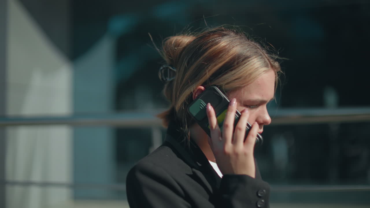 Young lady in business attire on phone call listens attentively while strolling home from work with focused expression, carrying handbag beside glass building reflecting cityscape