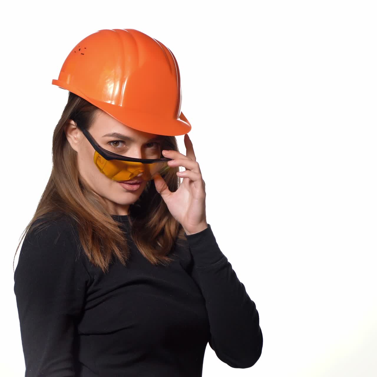 A joyful woman in orange helmet for building is trying on goggles and posing in it on a white background in the studio.