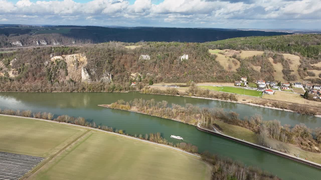 An aerial view of a boat on the Danube River, with a town, agricultural fields, and dramatic rocky cliffs in the background. A great view of the German landscape
