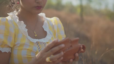 A-close-up-shot-of-an-Asian-female-holding-a-broken-clay-pot,-carefully-putting-the-pieces-together-of-the-fragile-sentimental-piece-of-pottery-while-sitting-in-a-grass-field-outdoors,-India