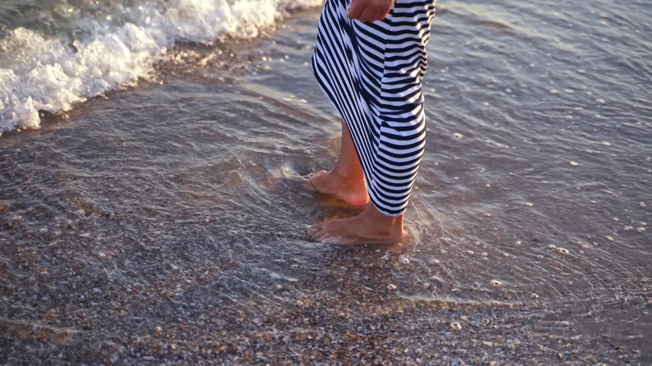 Woman playing with water on beach. Female legs in water. Woman in long dress splashing and playing water by feet on foamy waves background.