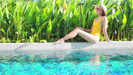 a full-body profile a young attractive woman on a one-piece bathing suit and sun hat sitting on the edge of a swimming pool taking in the sun