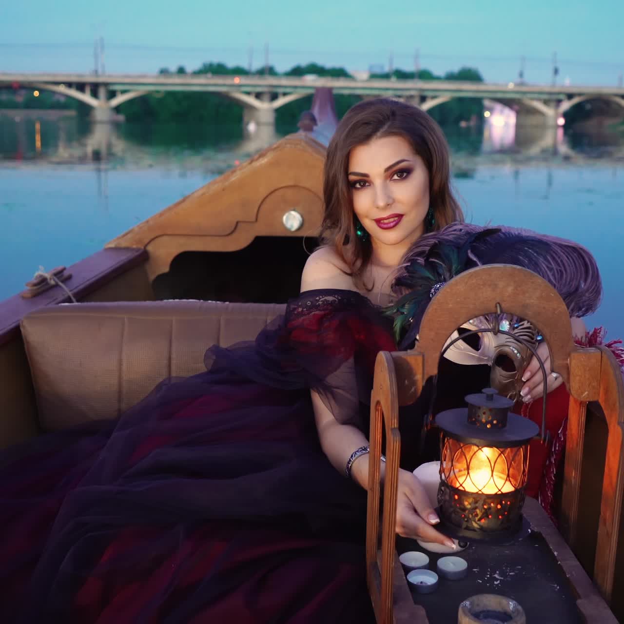 An elegant woman in a burgundy long dress is touching a decorative lantern and smiling on the gondola on the background of the river in the evening. Carnival