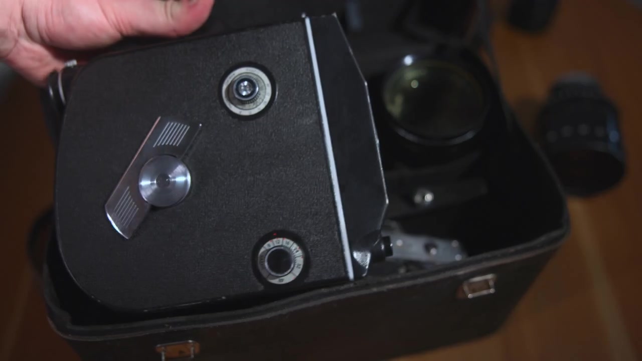 Close-up top view of an open black carrying case holding a vintage optical device with a visible lens and metal fittings, resting on a wooden surface under