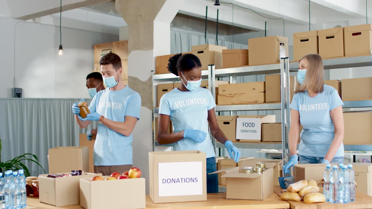 Multiethnic group of volunteers in facial mask packing boxes with food and clothes in charity warehouse