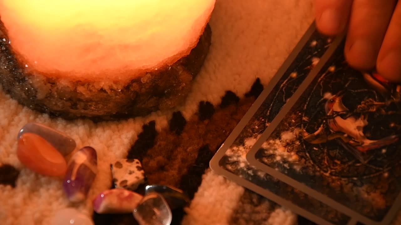 primer plano de la mano de un hombre tomando cartas del tarot una por una, cerca de algunas piedras y una lámpara de sal, sobre una alfombra