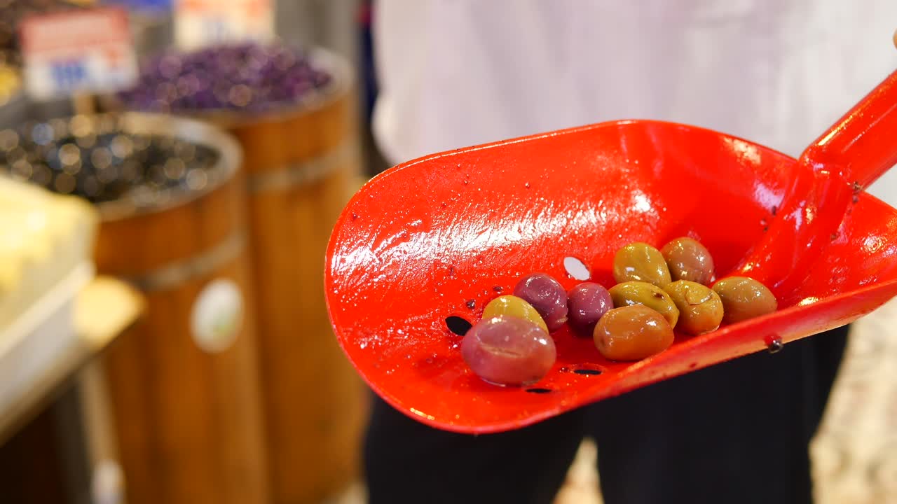 una persona que sostiene una cuchara roja con aceitunas verdes, negras y púrpuras. las aceitunas se exhiben para la venta en un mercado.