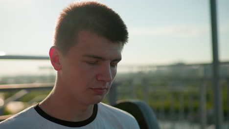 close up of man in white top looking downward with sunlight reflecting gently off his face, showing a calm and introspective expression, with background featuring greenery and iron railing