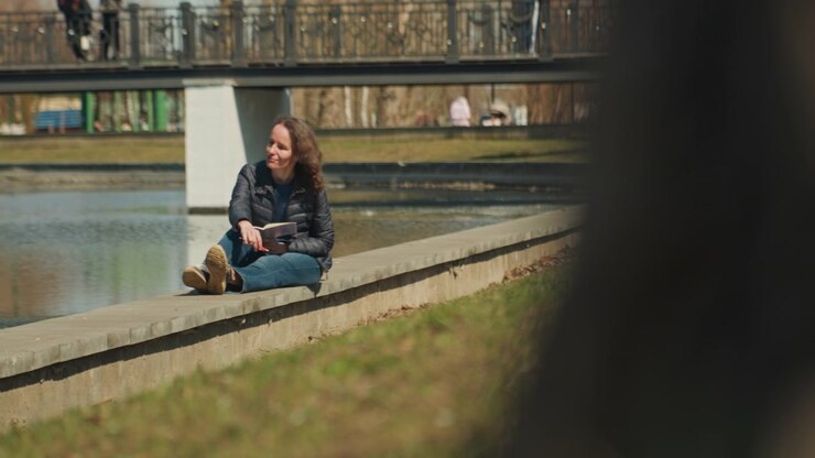 Close up of young woman reading book by calm water body in park setting holding pen while seated on concrete edge with bridge and people walking above in background. Soft daylight