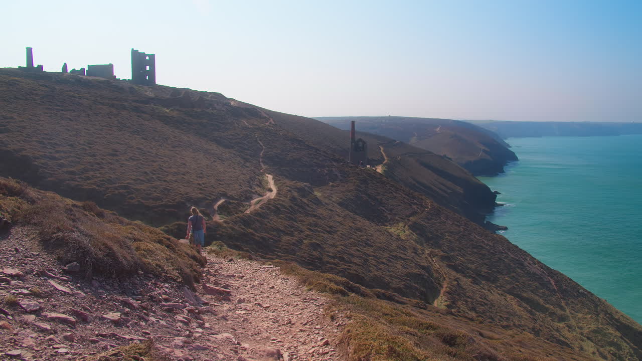 Travelers Walking Along The North Coast At Chapel Porth Beach Near Wheal Coates Tin Mine In United Kingdom. Wide Shot