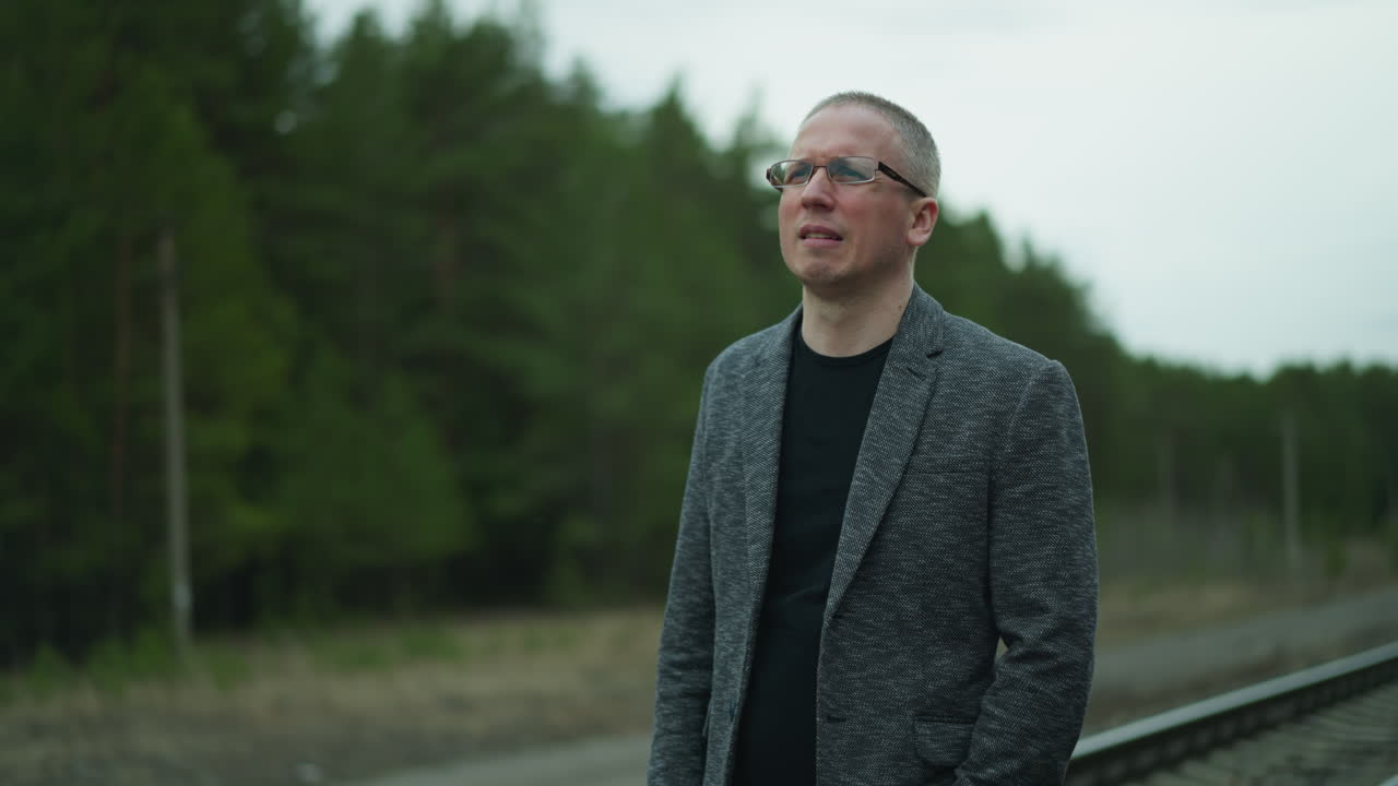 Close-up view of a man wearing a gray jacket standing on a railway track, putting on his glasses with one hand while looking thoughtfully into the distance, with a blurred background