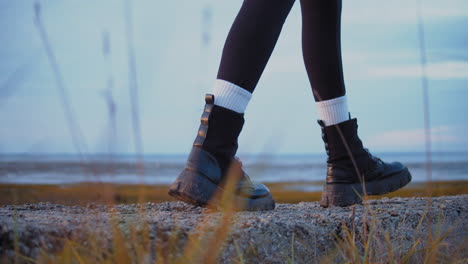 las piernas de un individuo con botas de guardabosques negras caminando lentamente por un camino costero, toma a nivel del suelo